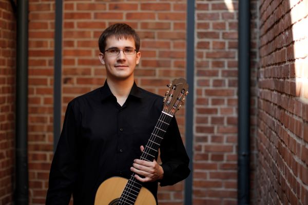 Młody mężczyzna w okularach ubrany w czarną koszulę. W lewej ręce trzyma gitarę klasyczną.